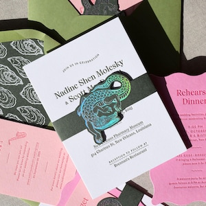 May include: A wedding invitation suite featuring white cards with black text, including the names Nadine Shen Molesky and Scott M, and a dark green belly band adorned with a holographic alligator cutout. Includes pink and olive green envelopes.