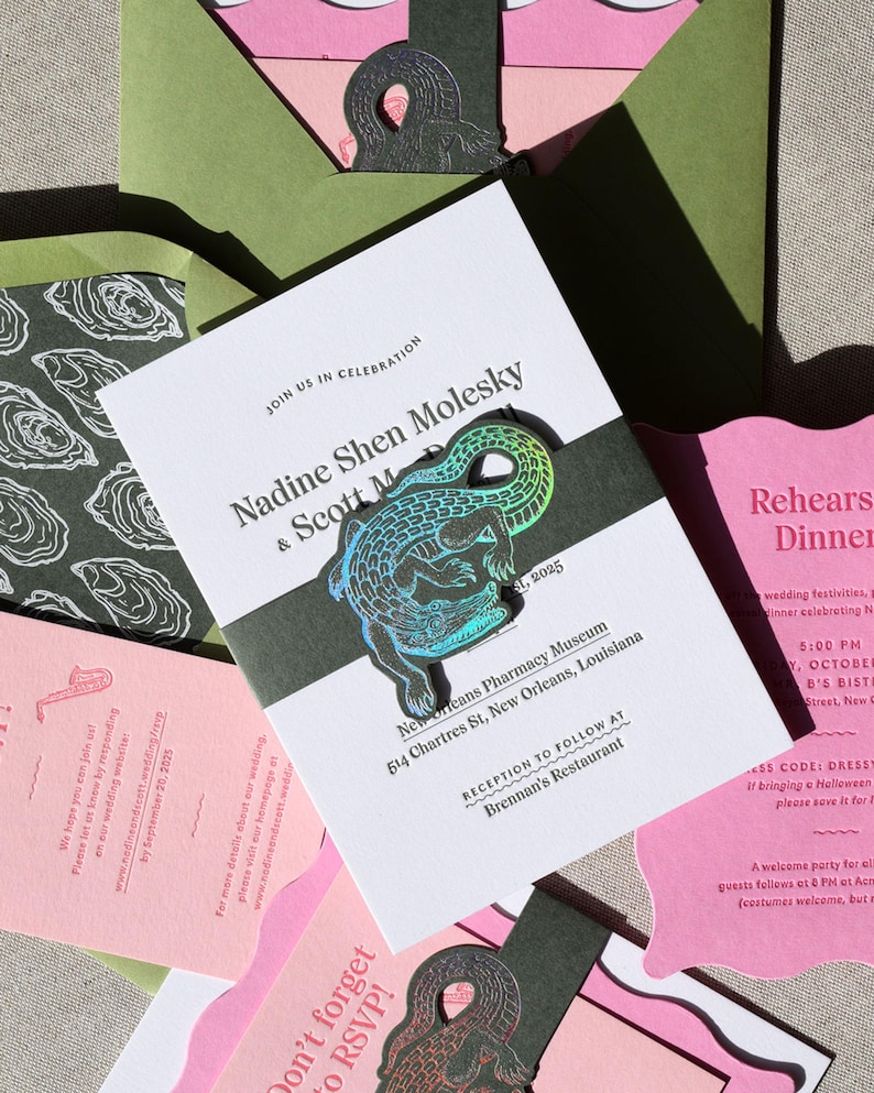 May include: A wedding invitation suite featuring white cards with black text, including the names Nadine Shen Molesky and Scott M, and a dark green belly band adorned with a holographic alligator cutout. Includes pink and olive green envelopes.