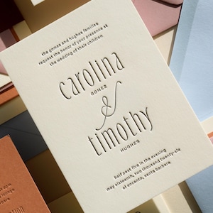 May include: An off-white wedding invitation card with debossed black text reading Carolina Gomez and Timothy Hughes, including the date May sixteenth, two thousand twenty-six, and location El Encanto, Santa Barbara, surrounded by colorful envelopes.