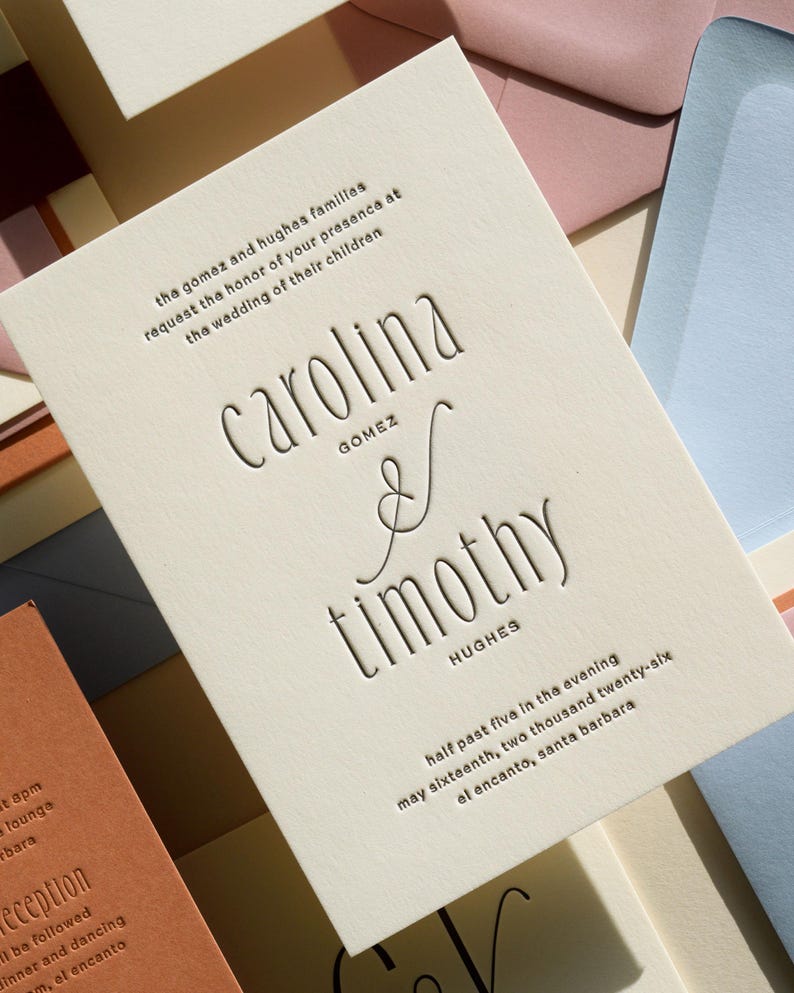 May include: An off-white wedding invitation card with debossed black text reading Carolina Gomez and Timothy Hughes, including the date May sixteenth, two thousand twenty-six, and location El Encanto, Santa Barbara, surrounded by colorful envelopes.