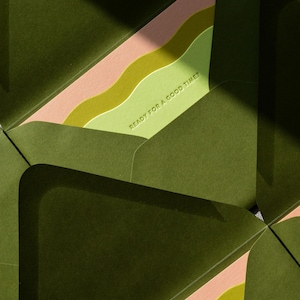 May include: A set of dark olive green envelopes with open flaps revealing a light green interior liner. The liner features wavy bands of pink and lime green, with the text READY FOR A GOOD TIME? embossed in a clean, sans-serif font.