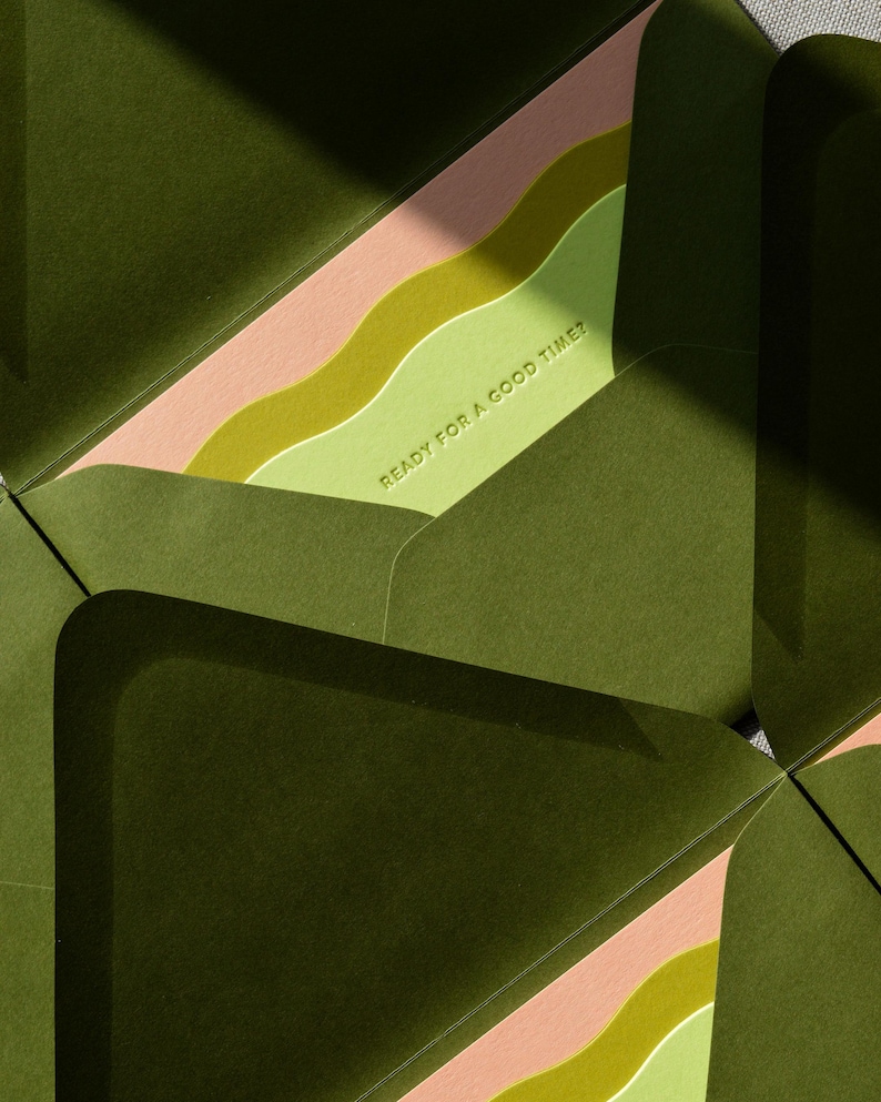 May include: A set of dark olive green envelopes with open flaps revealing a light green interior liner. The liner features wavy bands of pink and lime green, with the text READY FOR A GOOD TIME? embossed in a clean, sans-serif font.