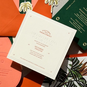 May include: A square wedding invitation card in cream with embossed text reading Together With Their Families Amanda Peyton & James Eno, December Twelfth, and event details. Surrounded by orange and dark green envelopes and palm tree tags.
