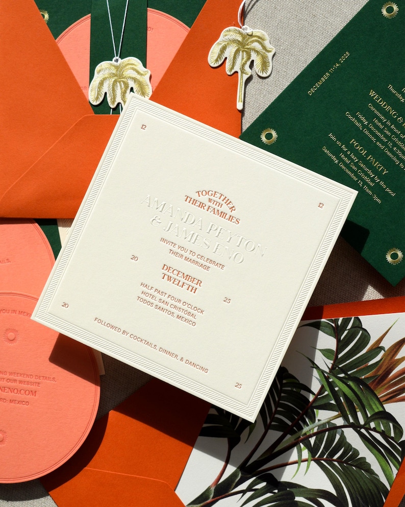 May include: A square wedding invitation card in cream with embossed text reading Together With Their Families Amanda Peyton & James Eno, December Twelfth, and event details. Surrounded by orange and dark green envelopes and palm tree tags.