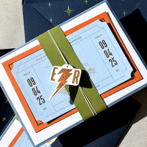 May include: A wedding invitation set featuring a light blue ticket-style card with the text ELLY & R and RSVP 09 04 25, layered over an orange card. An olive green paper band holds the set together, accented by a white tag with a copper lightning bolt and the letters E and R. The set is tucked into a dark blue envelope with a gold star pattern.