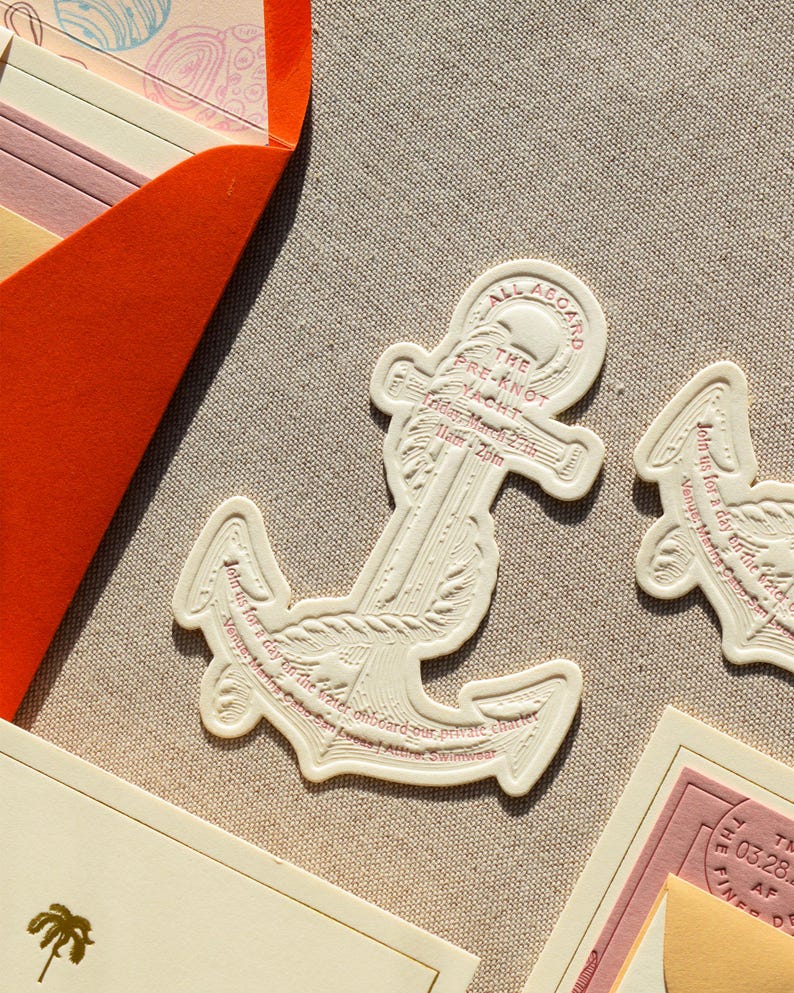 May include: An anchor-shaped invitation card in cream-colored cardstock with embossed details and red text. The text reads: ALL ABOARD THE PRE-KNOT YACHT, Friday March 27th, 11am-2pm, Join us for a day on the water onboard our private charter, Venue: Marina Cabo San Lucas | Attire: Swimwear.