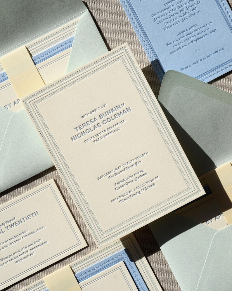 May include: A cream-colored wedding invitation suite featuring embossed dark text reading Teresa Bunkin and Nicholas Coleman, with matching light blue enclosure cards, envelopes, and decorative borders, all arranged in a layered, formal stationery set.