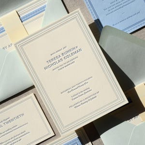 May include: A cream-colored wedding invitation suite featuring embossed dark text reading Teresa Bunkin and Nicholas Coleman, with matching light blue enclosure cards, envelopes, and decorative borders, all arranged in a layered, formal stationery set.