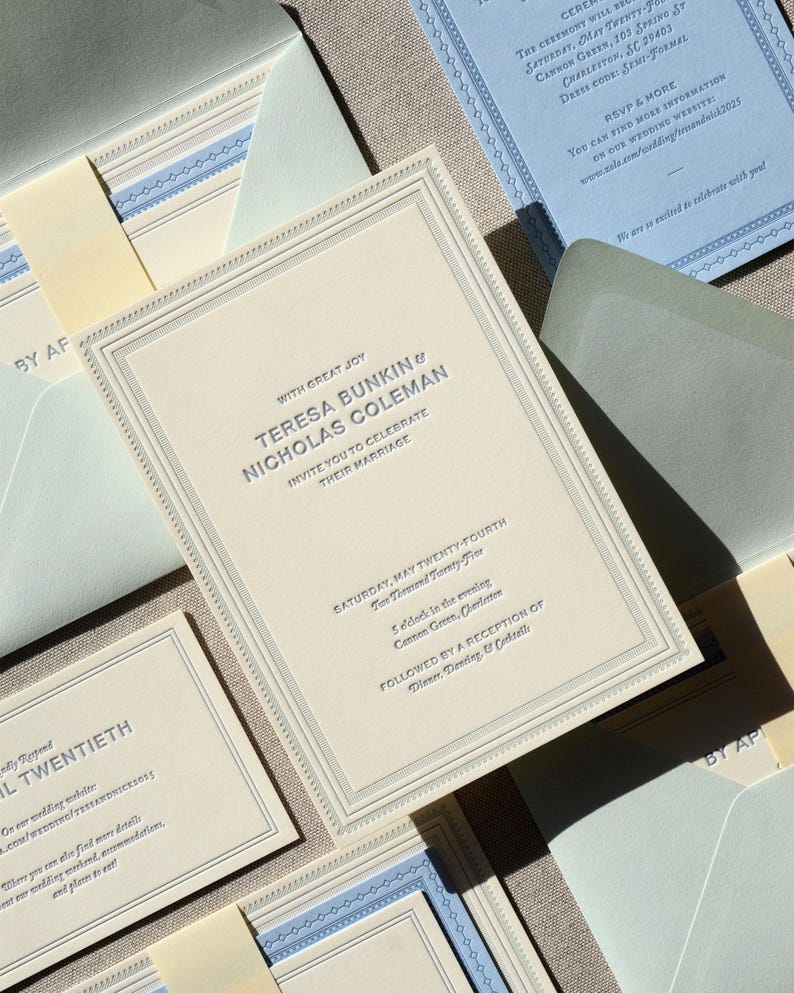 May include: A cream-colored wedding invitation suite featuring embossed dark text reading Teresa Bunkin and Nicholas Coleman, with matching light blue enclosure cards, envelopes, and decorative borders, all arranged in a layered, formal stationery set.