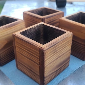 May include: Four square wooden planters with a natural wood grain finish. The planters are stained in varying shades of brown, with visible wood grain and a square opening at the top. They are arranged on a light blue surface.