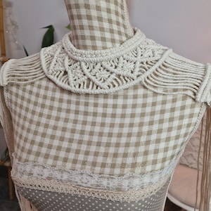 Puede incluir: Un collar de macramé blanco con detalles de flecos. El collar se muestra en un maniquí con un patrón de cuadros.