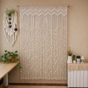 Puede incluir: Una cortina de macramé color crema cuelga de una barra de madera, con un diseño geométrico en la parte superior y detalles anudados. También se ve un tapiz de macramé con plantas y una mesa de madera con una planta en maceta.