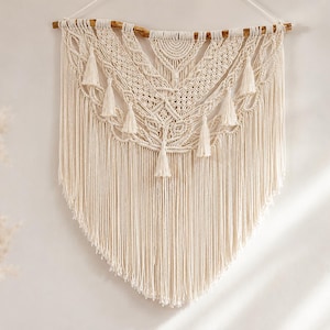 Puede incluir: Un tapiz de macramé de color crema colgado de una rama de madera natural. La pieza presenta intrincados patrones de nudos, un diseño en forma de V y múltiples borlas colgantes a lo largo del borde inferior, rematado con largos flecos.