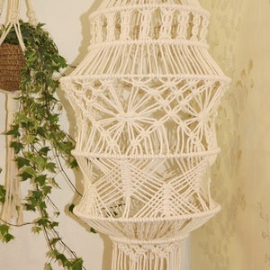 Puede incluir: Una lámpara colgante de macramé blanca con un diseño geométrico. La lámpara está hecha de cordón de algodón y tiene una franja en la parte inferior.