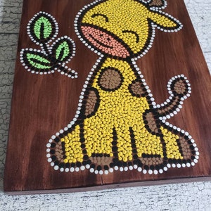 Puede incluir: Una placa de madera con un dise&ntilde;o de arte de puntos pintado a mano de una jirafa amarilla con manchas marrones. La jirafa sonr&iacute;e y tiene una hoja verde en la boca. El fondo es marr&oacute;n.