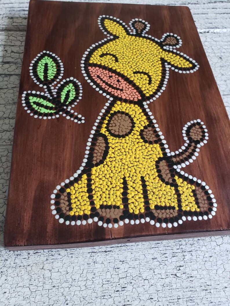 Puede incluir: Una placa de madera con un dise&ntilde;o de arte de puntos pintado a mano de una jirafa amarilla con manchas marrones. La jirafa sonr&iacute;e y tiene una hoja verde en la boca. El fondo es marr&oacute;n.