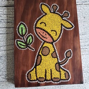 Puede incluir: Una placa de madera con una jirafa amarilla pintada en ella usando una t&eacute;cnica de pintura de puntos. La jirafa tiene manchas marrones y una nariz rosa. La jirafa est&aacute; sonriendo y tiene una hoja verde a su lado.