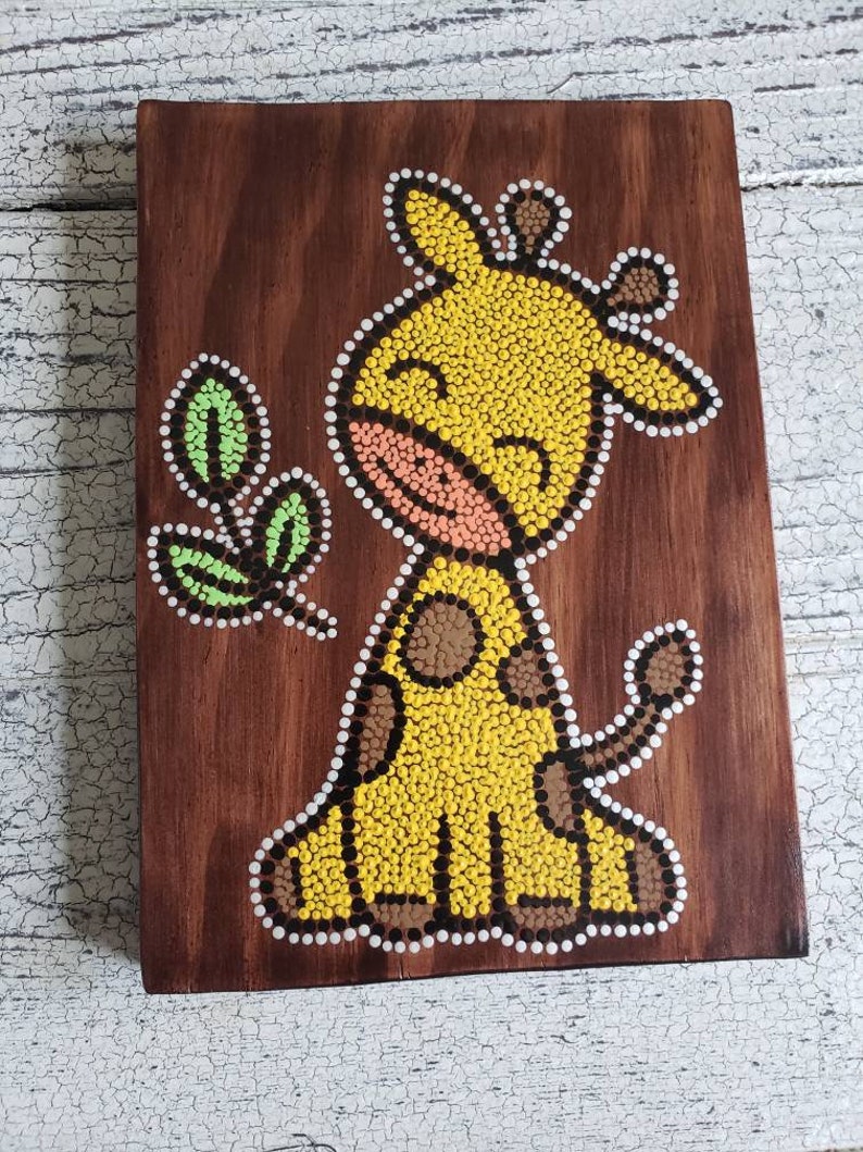 Puede incluir: Una placa de madera con una jirafa amarilla pintada en ella usando una t&eacute;cnica de pintura de puntos. La jirafa tiene manchas marrones y una nariz rosa. La jirafa est&aacute; sonriendo y tiene una hoja verde a su lado.