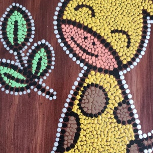 Puede incluir: Una pintura de puntos amarillos, marrones, negros, blancos, rosas y verdes sobre un fondo de madera marr&oacute;n. La pintura representa un p&aacute;jaro con una hoja en su pico.