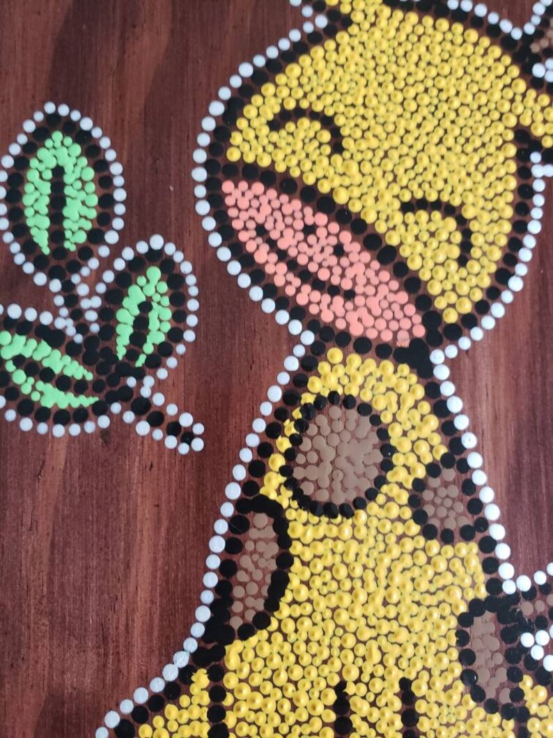 Puede incluir: Una pintura de puntos amarillos, marrones, negros, blancos, rosas y verdes sobre un fondo de madera marr&oacute;n. La pintura representa un p&aacute;jaro con una hoja en su pico.