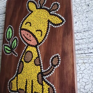 Puede incluir: Un lienzo de madera con una jirafa amarilla pintada en estilo de arte de puntos. La jirafa tiene manchas marrones y una nariz rosa. El fondo es de madera marr&oacute;n.