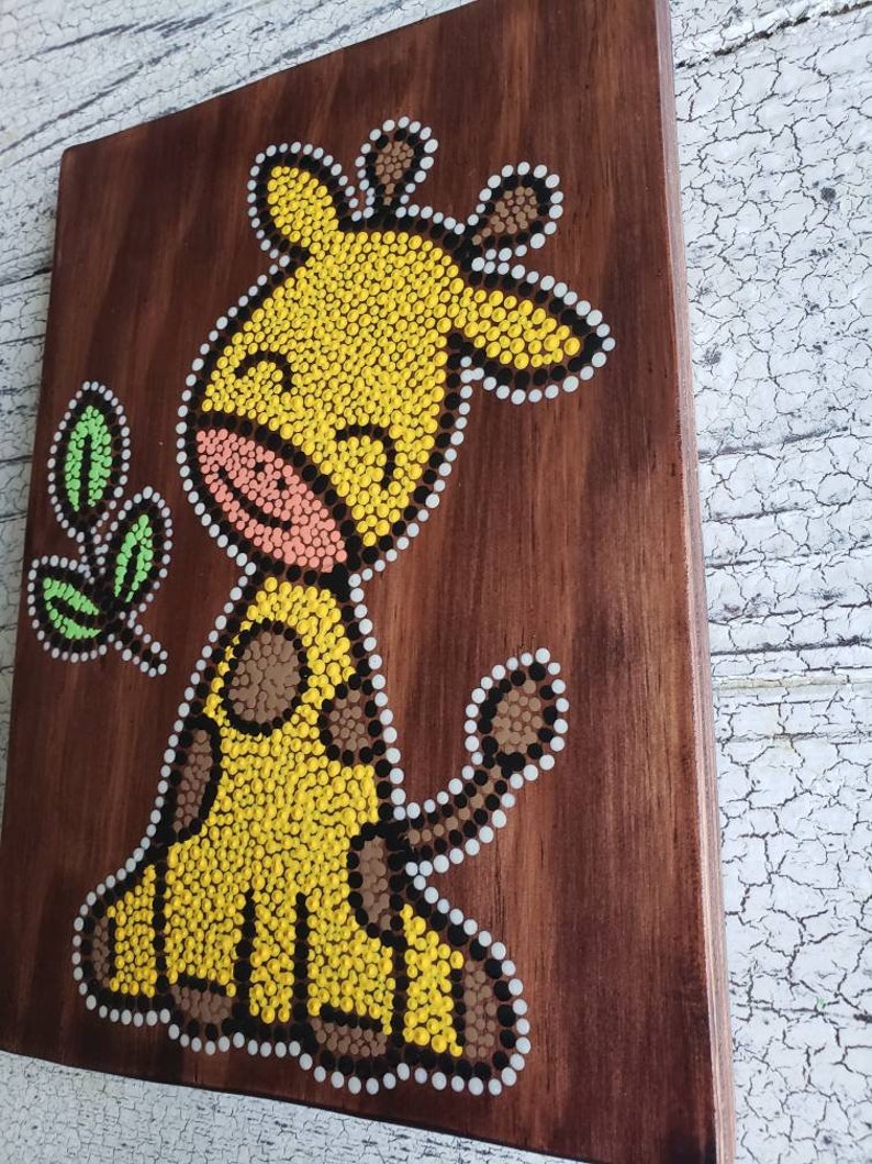Puede incluir: Un lienzo de madera con una jirafa amarilla pintada en estilo de arte de puntos. La jirafa tiene manchas marrones y una nariz rosa. El fondo es de madera marr&oacute;n.