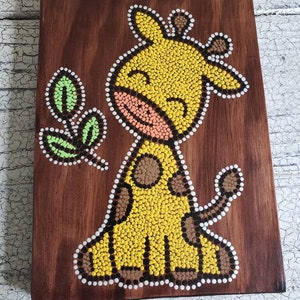 Puede incluir: Una placa de madera con un dise&ntilde;o de arte de puntos pintados a mano de una jirafa amarilla con manchas marrones y una hoja verde. La jirafa tiene una cara sonriente y est&aacute; pintada en un estilo caprichoso.