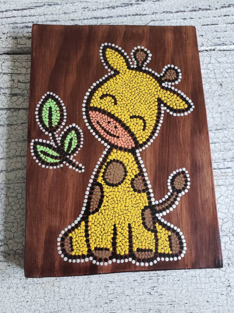 Puede incluir: Una placa de madera con un dise&ntilde;o de arte de puntos pintados a mano de una jirafa amarilla con manchas marrones y una hoja verde. La jirafa tiene una cara sonriente y est&aacute; pintada en un estilo caprichoso.