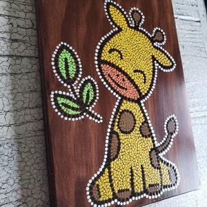 Puede incluir: Una placa de madera con una jirafa amarilla y una hoja verde pintadas con una t&eacute;cnica de pintura de puntos. La jirafa tiene manchas marrones y una boca rosa.