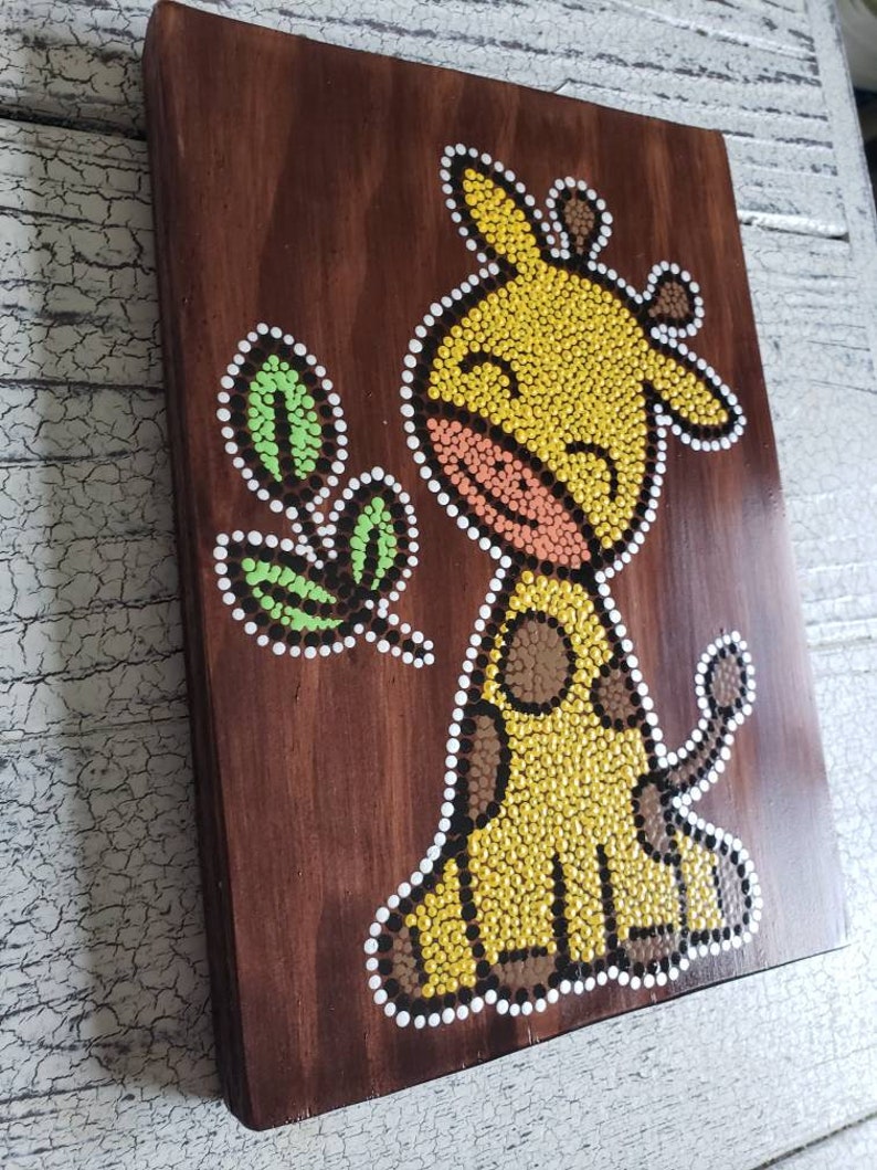 Puede incluir: Una placa de madera con una jirafa amarilla y una hoja verde pintadas con una t&eacute;cnica de pintura de puntos. La jirafa tiene manchas marrones y una boca rosa.