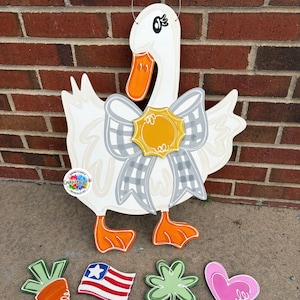 May include: A white wooden goose door hanger with an orange beak and feet, a gray plaid bow, and a yellow center. Includes interchangeable wooden cutouts of a carrot, American flag, shamrock, and heart.