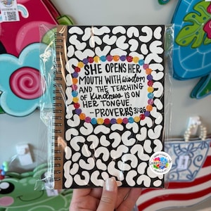 May include: A spiral-bound notebook with a black cover featuring a white abstract pattern. The notebook has the quote "She opens her mouth with wisdom and the teaching of kindness is on her tongue. Proverbs 31:26" in colorful text.