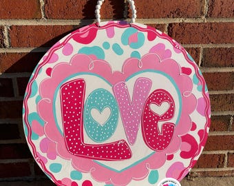 May include: Round wooden sign with the word "LOVE" in pink, white, and teal. The sign has a white background with pink and teal leopard print accents. A white beaded handle is attached at the top.