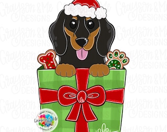 May include: A cartoon illustration of a black and tan dachshund wearing a Santa hat, peeking out of a green gift box tied with a red bow. The box contains gingerbread cookies. The text "Grayson & Me Designs" is visible.