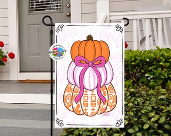 May include: A garden flag with a stacked pumpkin design. The top pumpkin is orange, the middle pumpkins are pink and white striped, and the bottom pumpkin is orange with a geometric pattern. A pink bow decorates the pumpkins.