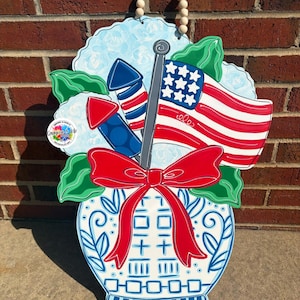 May include: Wooden patriotic door hanger featuring a blue and white vase with a red bow. The vase holds an American flag, fireworks, and green leaves. The background is light blue with white accents. The hanger has a beaded handle.