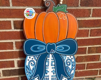 May include: Wooden pumpkin-shaped door hanger with an orange pumpkin design, a blue bow, and a vase with a blue and white pattern. The hanger has a string of wooden beads for hanging. The sign has the text "Creative Me".