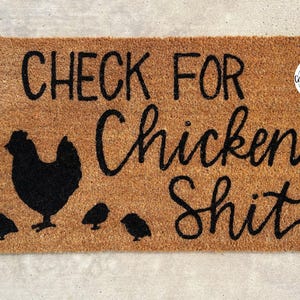 May include: A brown coir doormat with the text "CHECK FOR CHICKEN SHIT" in black cursive lettering. A black silhouette of a chicken and four chicks are also on the mat.