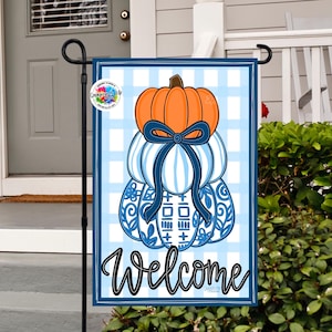 May include: A garden flag featuring a stacked pumpkin design with an orange top pumpkin, blue and white striped pumpkins, and a blue bow. The flag has a blue and white checkered background and the word "Welcome" in script.