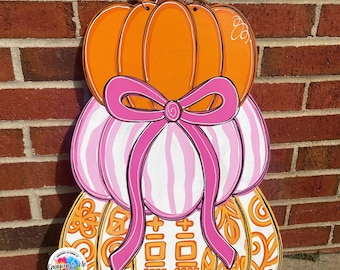 May include: A wooden pumpkin decoration with three stacked pumpkins. The top pumpkin is orange, the middle is pink and white striped, and the bottom is orange with a decorative pattern. A pink bow adorns the middle pumpkin, and a beaded handle is at the top.