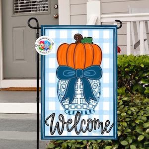 May include: A decorative garden flag featuring a pumpkin design. The pumpkin is orange with a brown stem and green leaves, set atop a blue and white vase with a blue bow. The word "Welcome" is written in black script.