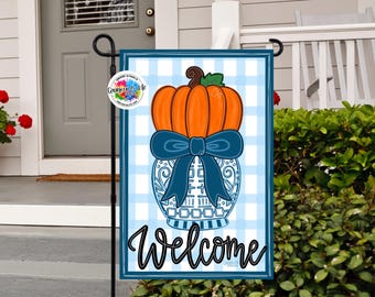May include: A decorative garden flag featuring a pumpkin design. The pumpkin is orange with a brown stem and green leaves, set atop a blue and white vase with a blue bow. The word "Welcome" is written in black script.