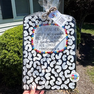 May include: A black and white decorative cutting board with a leopard print design. The board features the quote "She opens her mouth with wisdom and the teaching of kindness is on her tongue - Proverbs 31:26". The board is wrapped in cellophane.
