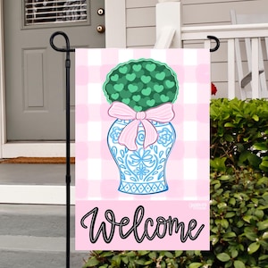 May include: A pink and white gingham garden flag with a blue and white vase with a green bush with heart-shaped leaves and a pink bow. The text "Welcome" is written in black cursive on the flag.