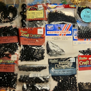 May include: A collection of black craft beads in various sizes and shapes, including faceted and round beads. Packaged in clear plastic bags and labeled with brand names like "Darice Craft Designer" and "Westrim Crafts."