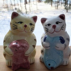 May include: Two ceramic cat salt and pepper shakers. The cat on the left is yellow with brown stripes and is holding a pink fish. The cat on the right is white with gray stripes and is holding a blue fish.