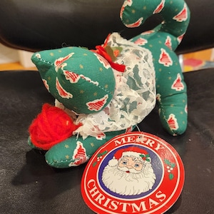 May include: A green fabric cat-shaped Christmas ornament with a red and white tree pattern. The cat has a white lace collar and a red yarn ball. A round tag with Santa Claus and "MERRY CHRISTMAS" is attached.