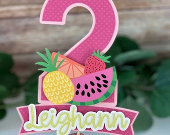 May include: Pink cake topper with a number 2, a pineapple, watermelon, and strawberry. The topper also has a pink banner with the name Leighann in gold glitter letters.