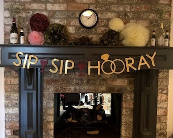 May include: A brick fireplace mantel decorated with a gold banner that reads "Sip Sip Hooray" with two gold rings. The mantel is decorated with wine bottles, pink and yellow tissue paper pom-poms, and greenery.