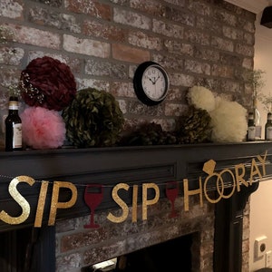 Puede incluir: Una pancarta de purpurina dorada con el texto "Sip Sip Hooray" y un anillo dorado con un diamante. La pancarta est&aacute; colgada sobre una chimenea con un marco de ladrillo. Tambi&eacute;n hay algunas bolas de papel decorativas en el fondo.